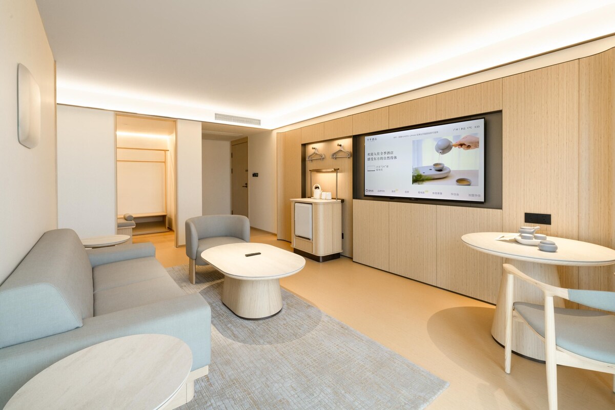 Ein modernes Hotelzimmer mit einer Couch, einem kleinen runden Tisch, einem an der Wand befestigten Fernseher und einem Holzschrank.