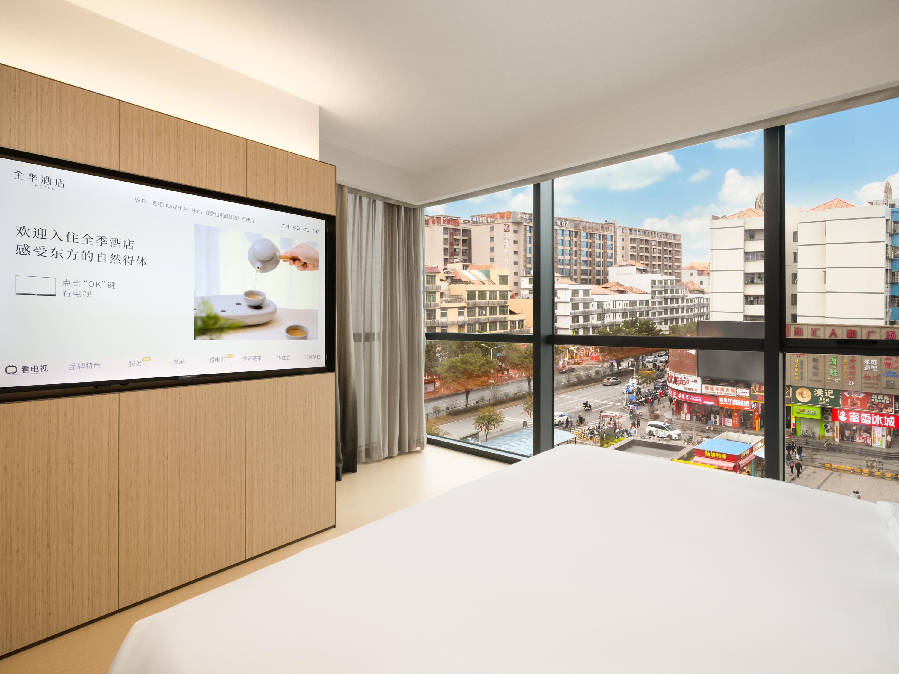 A hotel room with a large window, a television screen, and a bed.