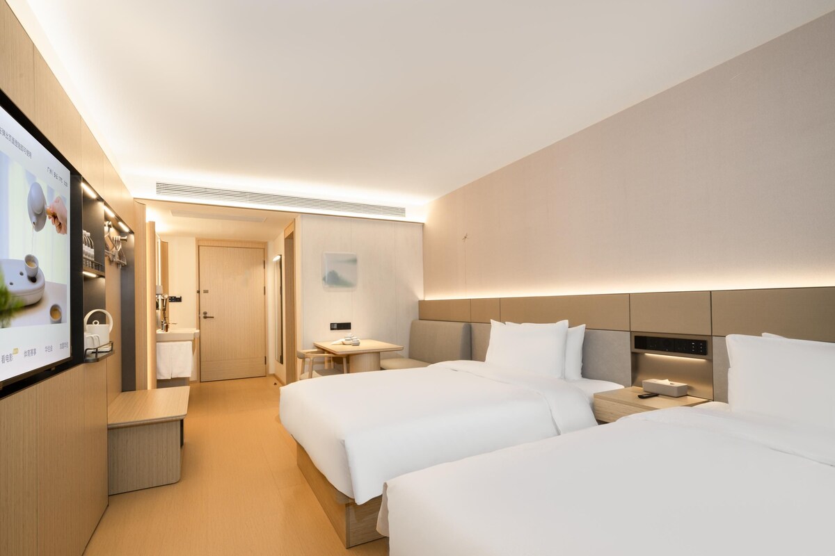 Ein Hotelzimmer mit zwei Betten, einem kleinen Tisch, einem Nachttisch, einem Fernseher und einem durch eine offene Tür einsehbaren Badezimmer.