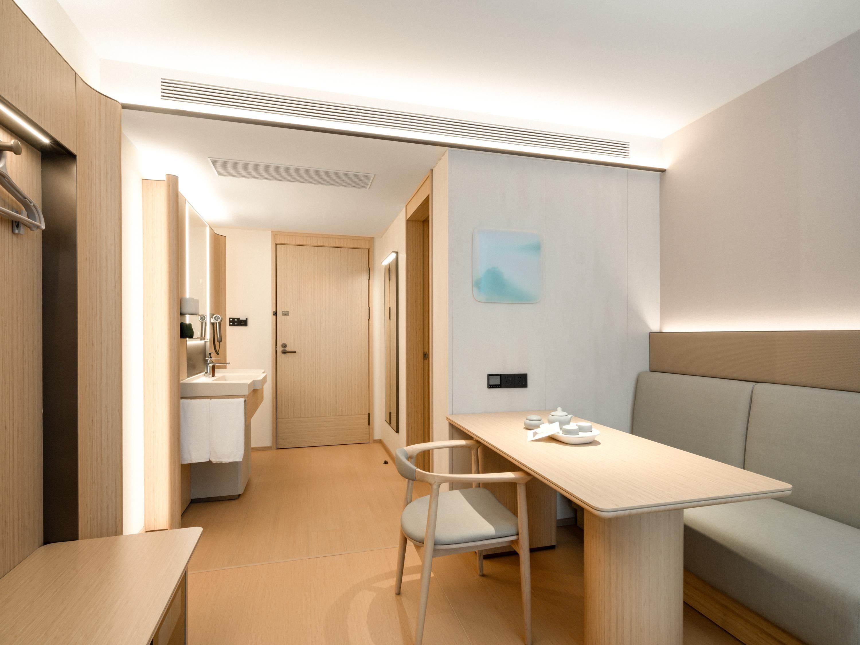 A modern hotel room with a wooden floor, a desk with a chair, a sofa, a bathroom with a sink and mirror, and a door leading to another room.