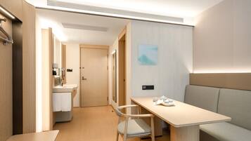A modern hotel room with a wooden floor, a desk with a chair, a sofa, a bathroom with a sink and mirror, and a door leading to another room.