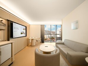 A modern hotel room with a flat-screen TV, a sofa, a dining table, and a view of the city through large windows.