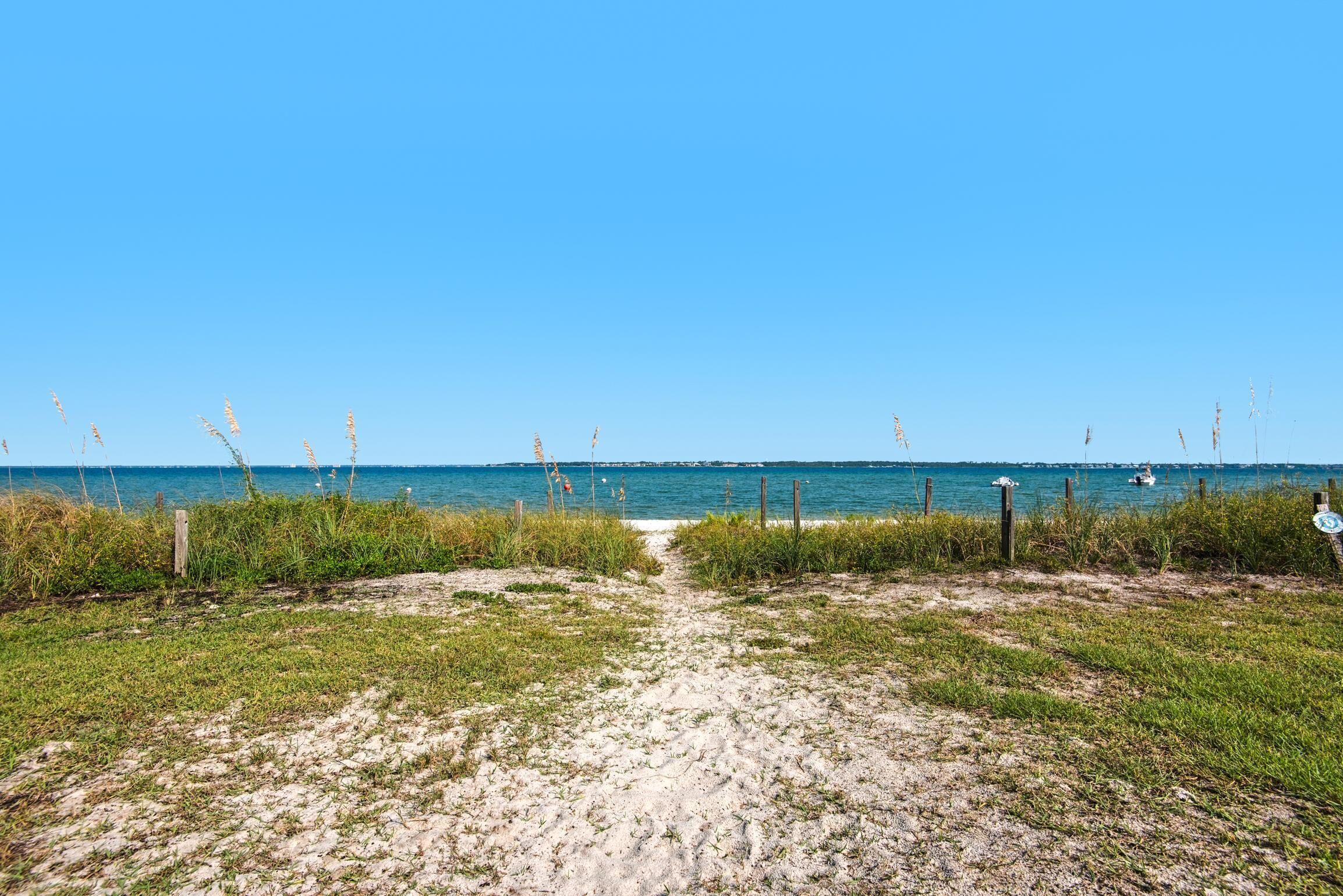 Un sentier de sable qui longe la plage et mène à l’océan, avec une végétation clairsemée et un ciel bleu dégagé.