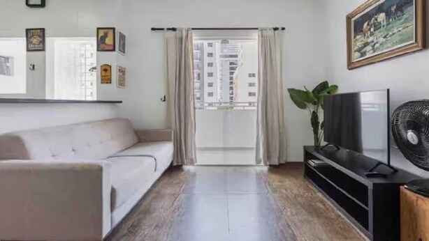 A living room with a sofa, a TV, a fan, and a view of buildings through a glass door. - Cozy apartment in the best location in Itaim Bibi. (São Paulo)