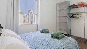 A hotel room with a bed, a window with a city view, a metal wardrobe, and a small table. - Cozy apartment in the best location in Itaim Bibi. (São Paulo)