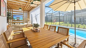 A wooden dining table with wicker chairs, a large umbrella, and a view of a swimming pool through a glass enclosure. - 2-Story Slide! Mickey & Minnie Clubhouse! MARIO Galaxy! Minion Room! (Clermont)