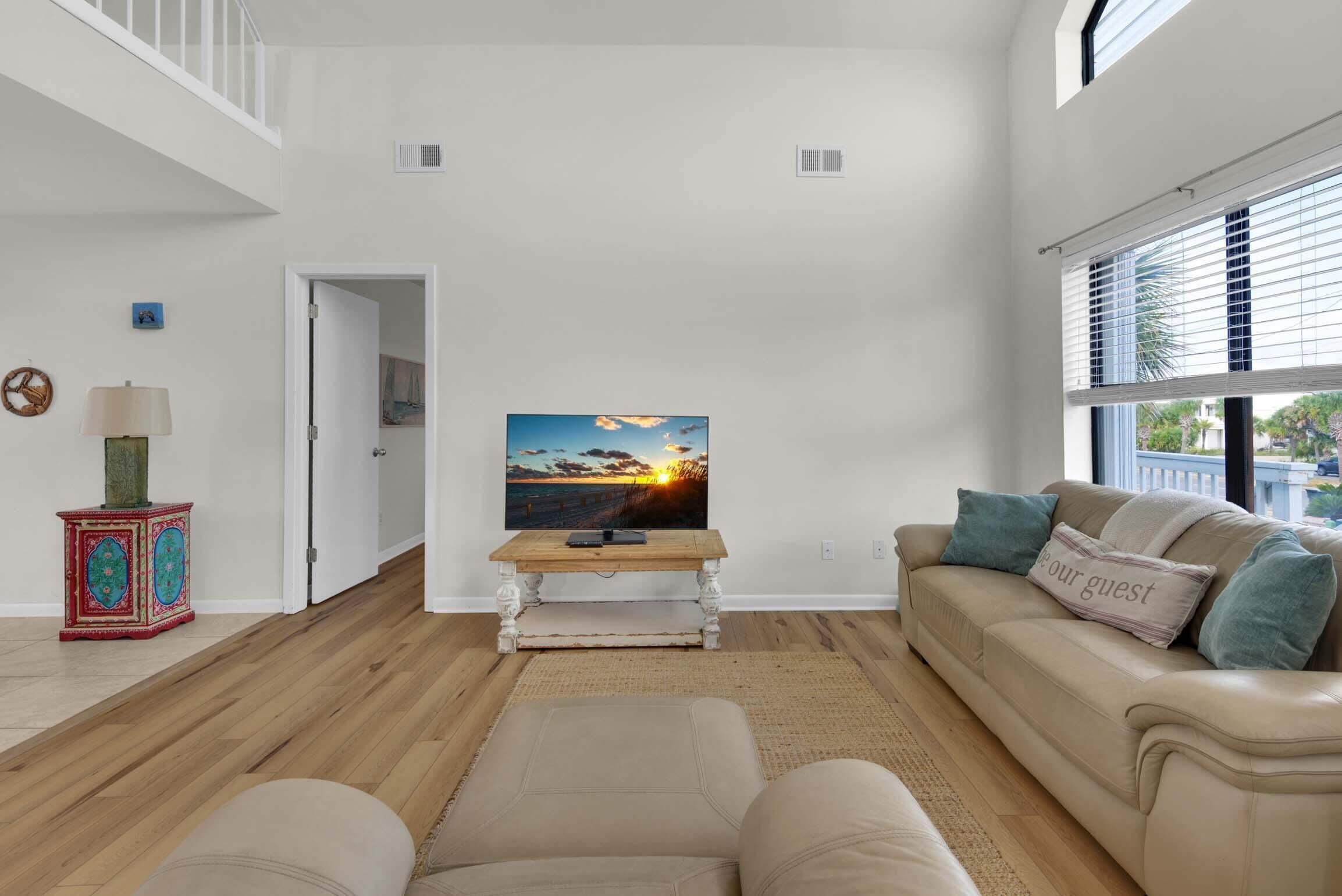 Ein Wohnzimmer mit Holzboden, einem beigen Sofa, einem Fernseher auf einem Holzschrank und einer Tür zu einem anderen Raum.