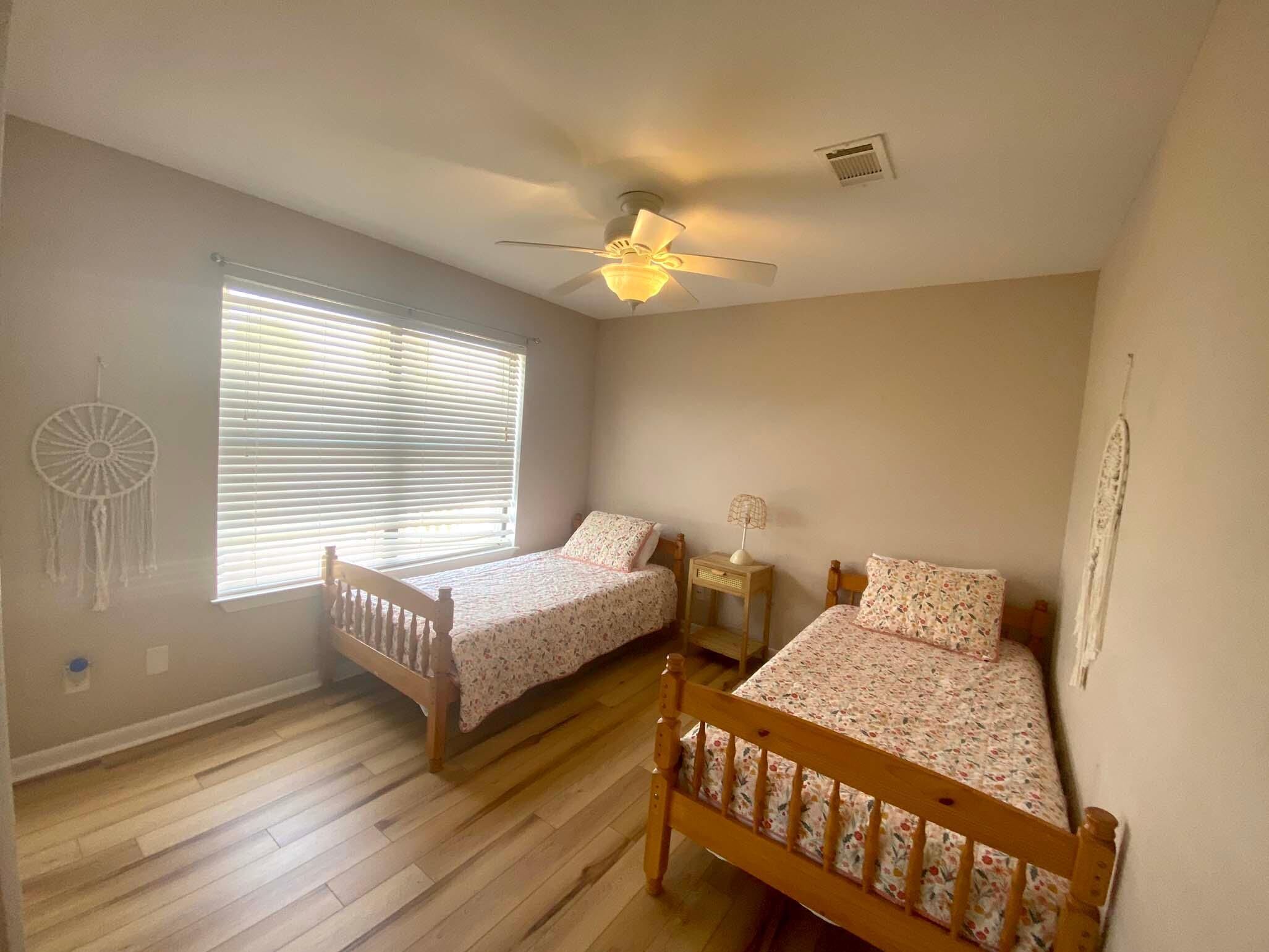 Un dormitorio con dos camas individuales, un ventilador de techo, una ventana con persianas y un atrapasueños en la pared.