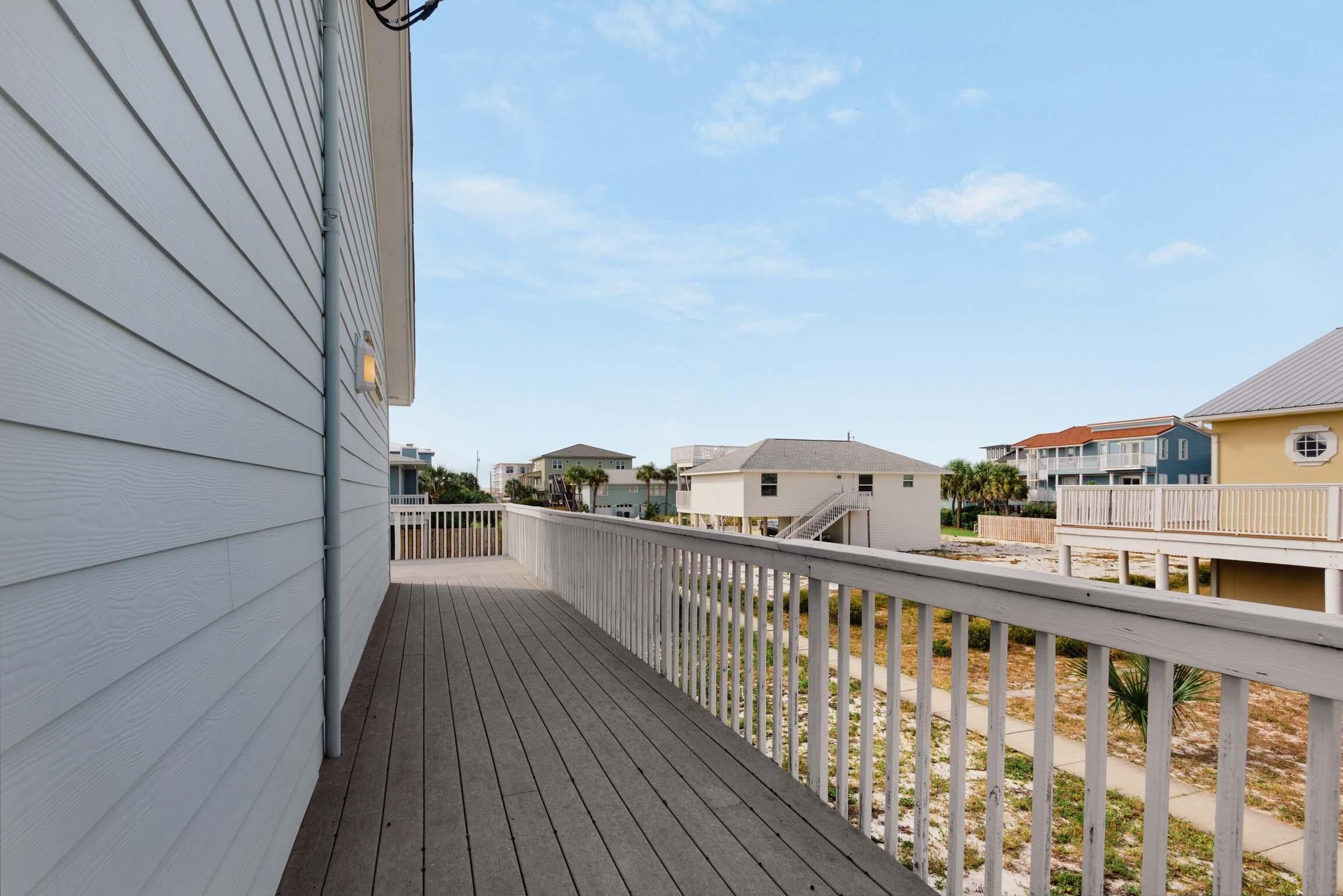 Une terrasse en bois avec une balustrade blanche, qui mène à une zone sablonne où l’on aperçoit des bâtiments en arrière-plan.