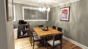 A dining area with a wooden table and chairs, a chandelier, a window with blinds, and a framed picture on the wall.