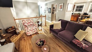 A living room with a wooden floor, a purple sofa, a patterned armchair, a small round table, and a staircase.
