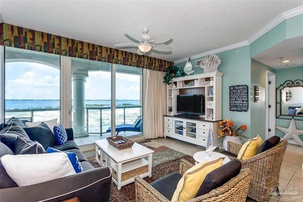 A spacious living room with a large TV, comfortable seating, a balcony with a view, and a ceiling fan. - Portofino Island Resort 3-706 (Pensacola Beach)