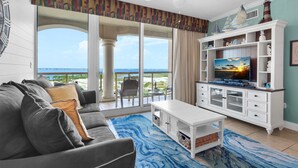 A living room with a gray sofa, a white coffee table, and a TV on a white cabinet. There is a large window with a view of the ocean. - Portofino Island Resort 3-706 (Pensacola Beach)