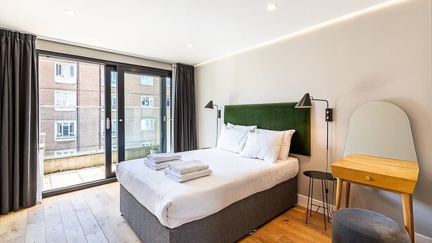 A modern hotel room with a large bed, a bedside table, a mirror, and a view of the city through a large window.