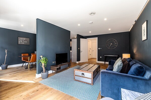 A modern living room with a dark blue wall, a blue sofa, a wooden coffee table, and a television on a stand.