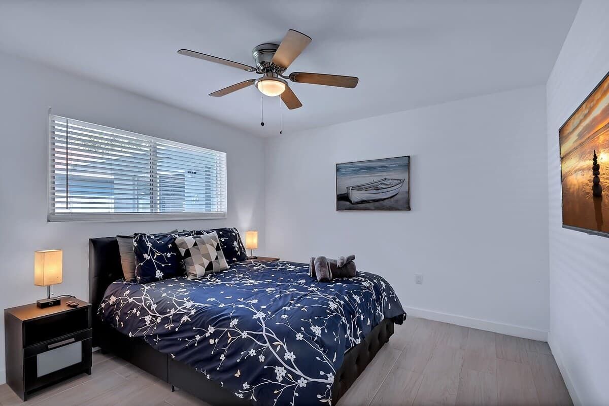 A bedroom with a bed, nightstands, a ceiling fan, a window with blinds, and a painting on the wall.