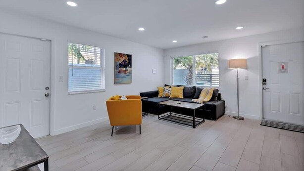 A modern living room with a sofa, armchair, and coffee table. There are two windows with blinds, a floor lamp, and a door with a handle. - Stylish 2BR Retreat in Miami's Design District! (Miami)