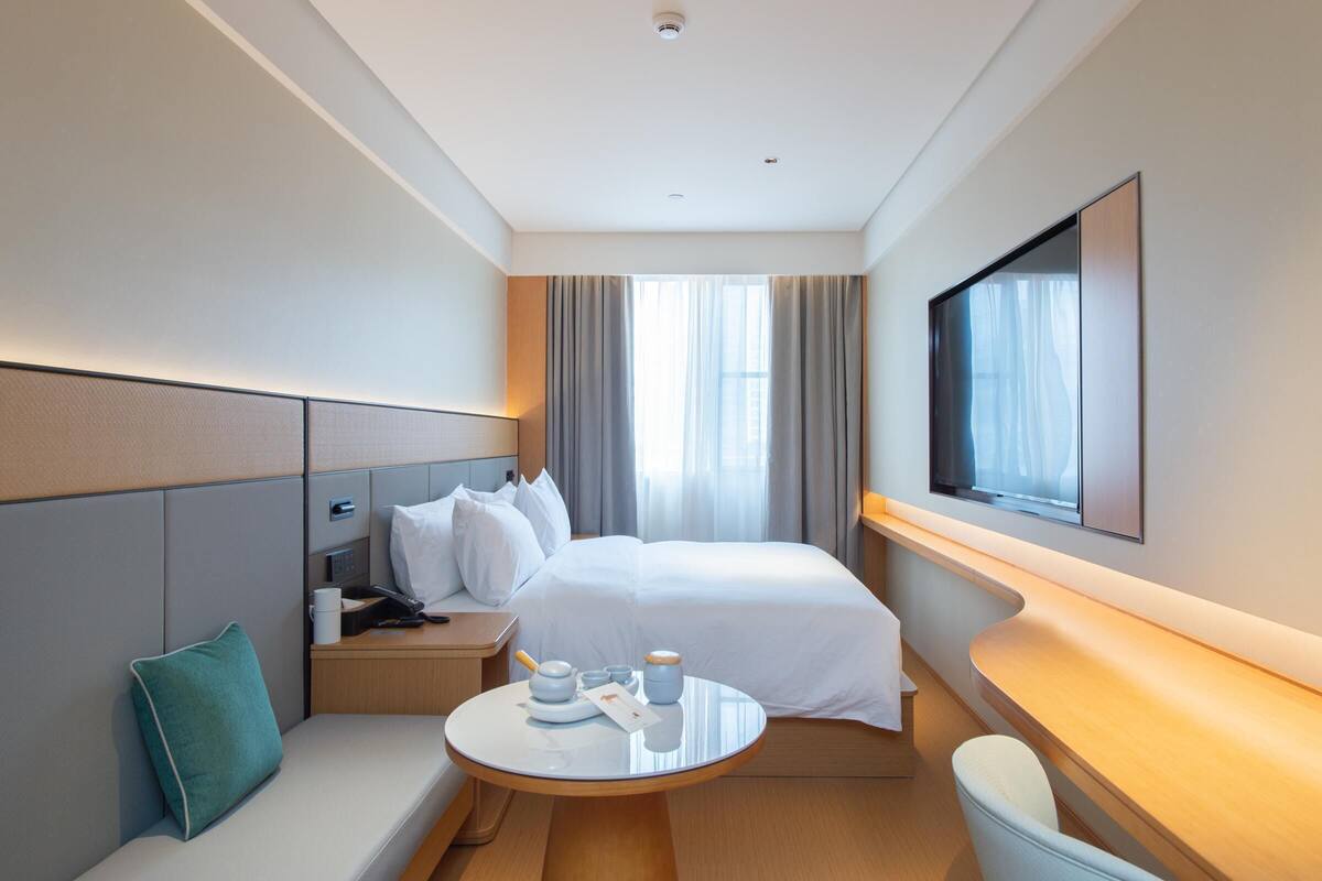 Een moderne hotelkamer met een groot bed, een nachtkastje met lamp, een klein rond tafeltje met kop en schotel, een bank met kussen, en een tv aan de muur.