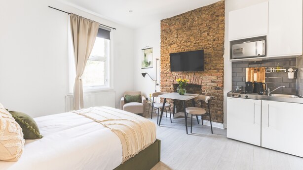 A modern bedroom with a large bed, a dining area with a table and chairs, a kitchen with white cabinets and a brick accent wall, and a flat-screen TV mounted above the counter. - Stylish & Petit Studio flat w/great tube connectio (Greater London)