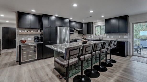A modern kitchen with dark cabinets, a large island with bar stools, and a view to the outdoors. - Cozy Haven - 4 En-Suite BR + Hot Tub + Fire Pit (Lead)