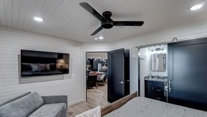A modern bedroom with a bed, a gray sofa, a ceiling fan, and a TV mounted on the wall. - Cozy Haven - 4 En-Suite BR + Hot Tub + Fire Pit (Lead)