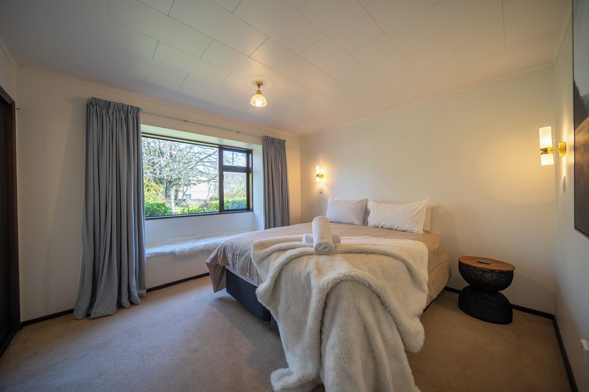 Ein Schlafzimmer mit einem Bett, einem Fenster, Vorhängen, einer Wandleuchte und einem kleinen runden Tisch.