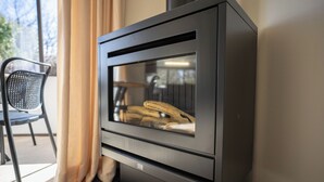 Een moderne houtkachel met glazen deur, in een kamer met een stoel en gordijnen.