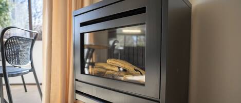 A modern wood-burning stove with a glass door, located in a room with a chair and curtains.