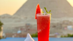 A tall glass of red beverage with a slice of watermelon and mint garnish, placed on a table with a view of a pyramid in the background. - Vysta pyramids hotel Cairo 
View pyramids 
renovated rooms
Restaurant with view (Giza Governorate)