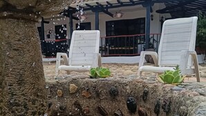 Uma fonte de pedra com água a jorrar, rodeada por pedras e plantas verdes. Duas cadeiras de descanso brancas estão posicionadas perto.