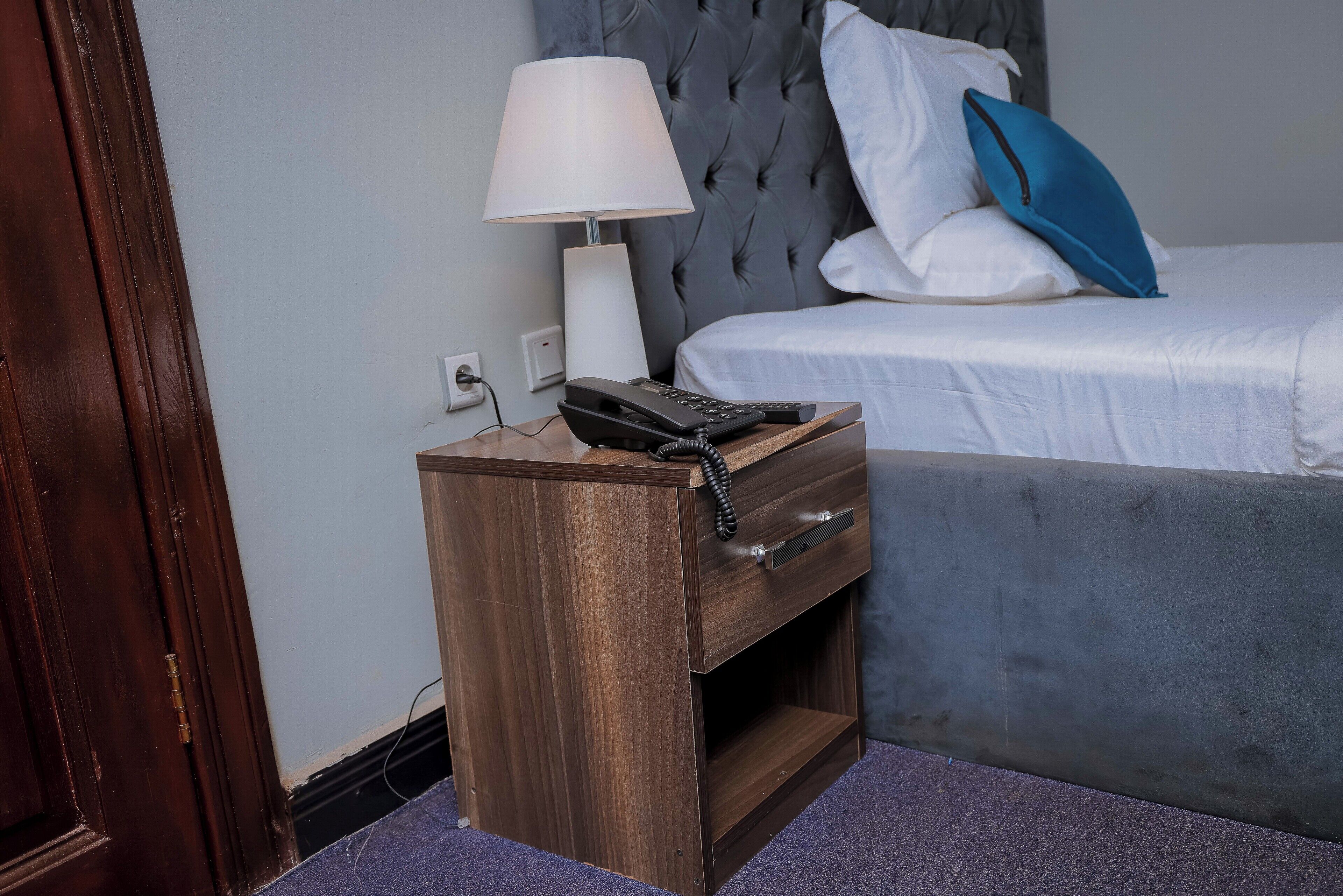 Ein Nachttisch aus Holz mit einer Lampe, einem Telefon und einer Schublade. Im Hintergrund ist ein Bett mit weißer Bettwäsche und einem blauen Kissen zu sehen.