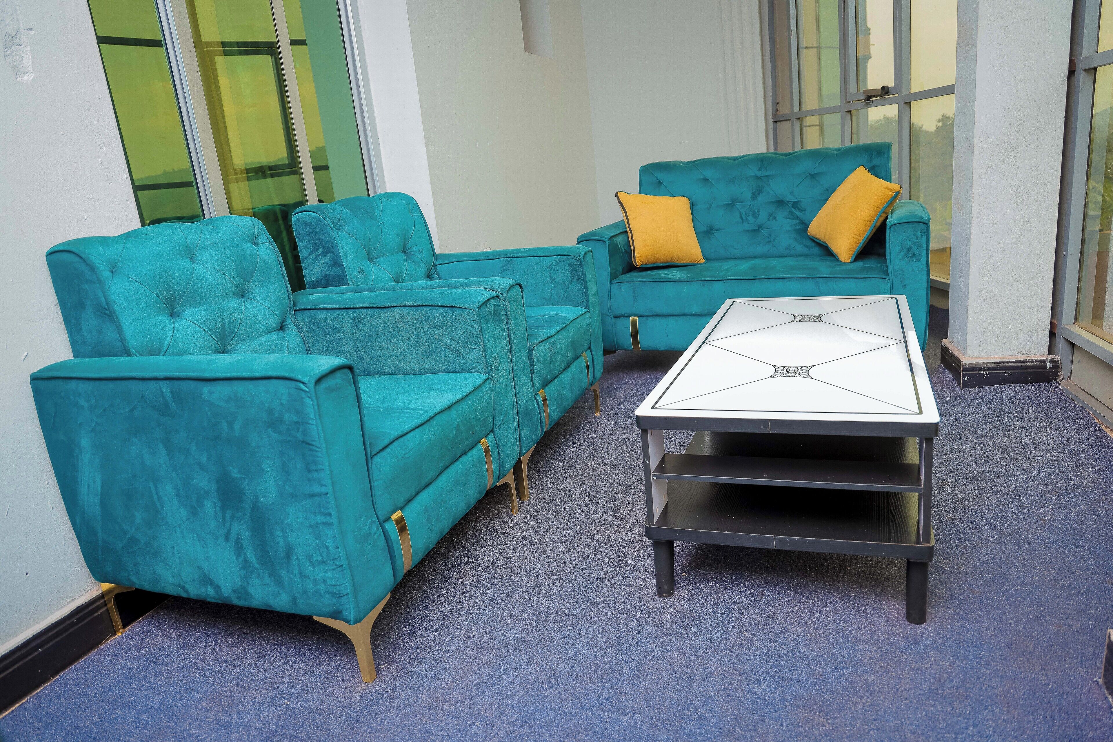 Un ensemble de canapés de couleur turquoise avec des coussins jaunes, une table basse blanche et un sol recouvert d’une moquette bleue, dans une pièce dotée de grandes fenêtres.