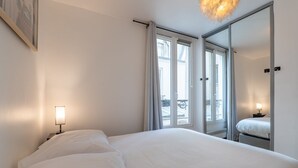 A hotel room with a large bed, two bedside lamps, a mirror, and a view of the outside balcony. - Cozy Hideaway in a Quiet Paris Passage (Paris)
