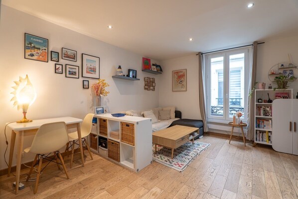 A cozy living room with a white sofa, a small wooden table, a desk with a lamp, and a bookshelf.