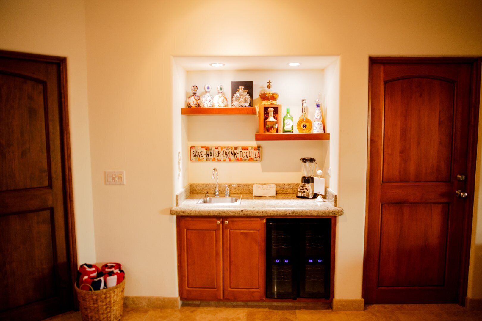 A compact kitchen with wooden cabinets, a granite countertop, a built-in dishwasher, and a shelf with various bottles and a sign that reads "SAVE-WATER-DRINK-TCQUILA".