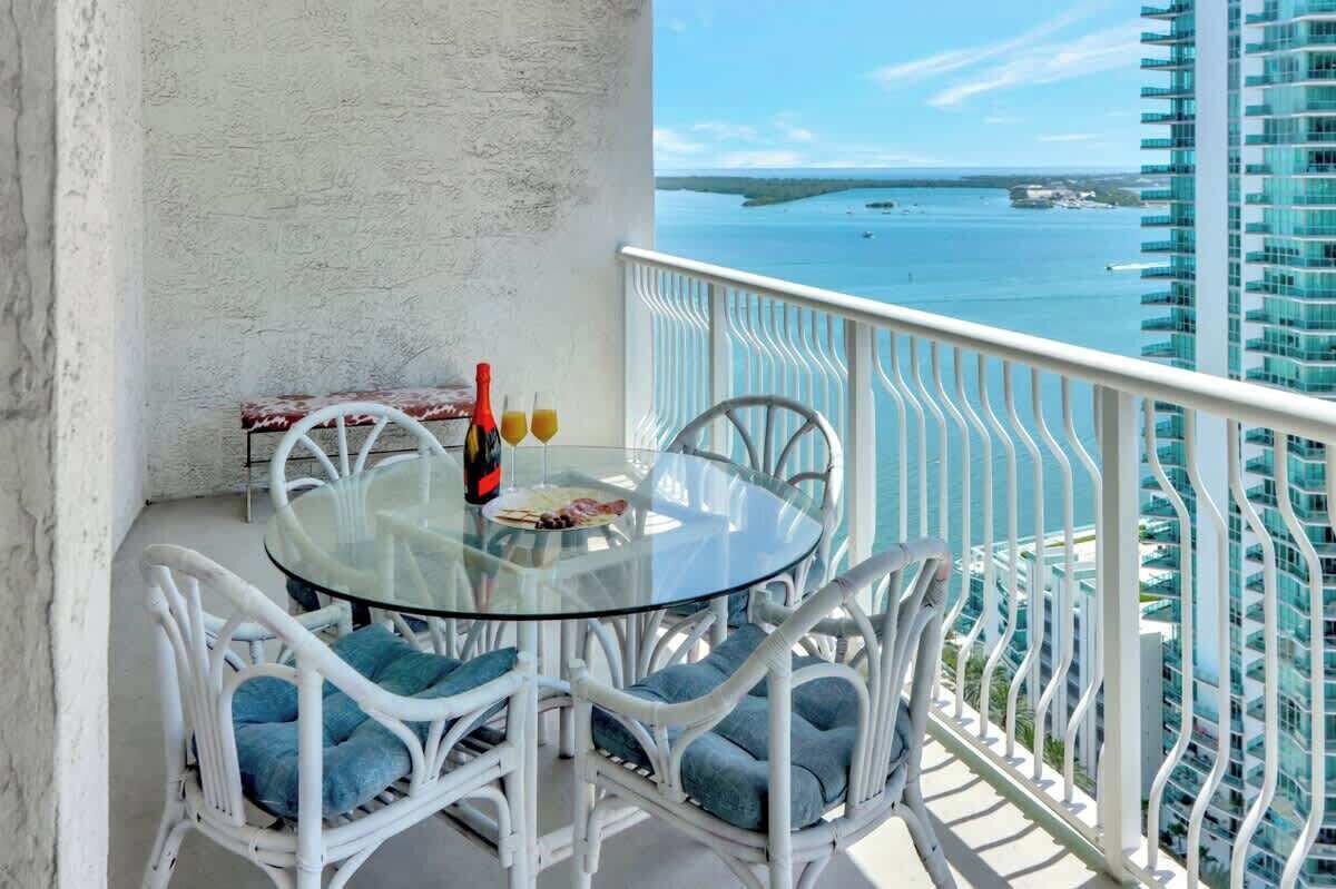 Ein Balkon mit Glastisch und Korbstühlen, der auf ein Gewässer und ein Hochhaus blickt.