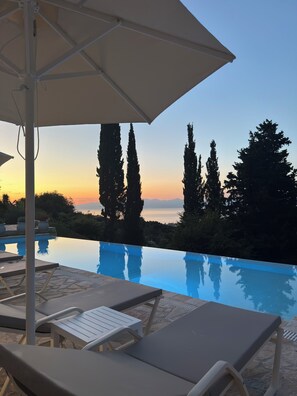 Une piscine à débordement avec des chaises longues et un parasol, offrant une vue sur un coucher de soleil, avec de grands arbres et un paysage lointain.