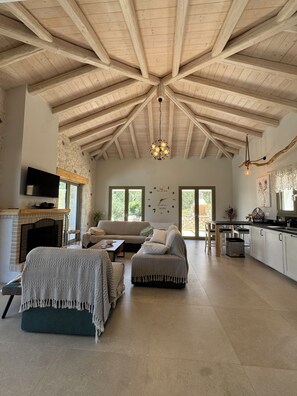 A spacious living area with a high wooden ceiling, a fireplace, a sofa, a coffee table, and a kitchen area with a dining table and chairs.