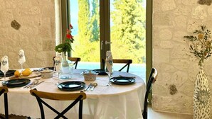 A dining table set for four with a white tablecloth, surrounded by stone walls and a large window with a view of trees.