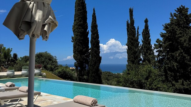 A poolside area with lounge chairs and an umbrella, overlooking a clear blue sky and a body of water.