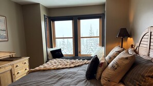 Une chambre avec un grand lit, une fenêtre donnant sur des arbres couverts de neige, une commode en bois et une lampe.