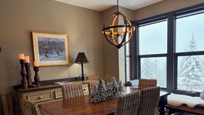 Un coin repas avec une table en bois, des chaises en osier, un lustre, un tableau encadré et une vue sur des arbres couverts de neige, visible à travers de grandes fenêtres.