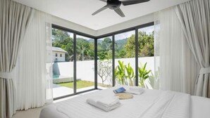 A modern bedroom with a large bed, a ceiling fan, and a view of greenery and a building outside.