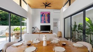 A modern dining area with a wooden table set for four, surrounded by white cushioned chairs, a large painting on the wall, and a television in the background.