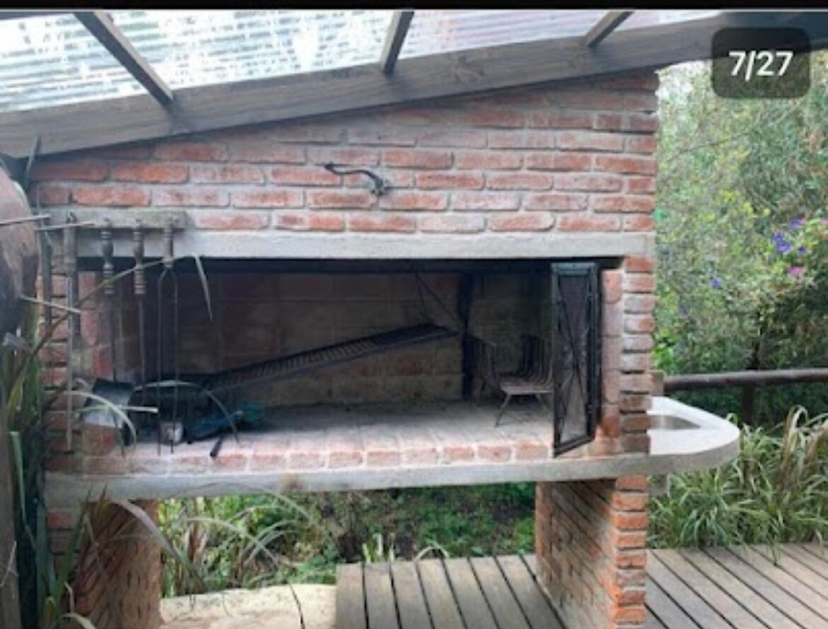 Ett utomhusområde med grillplats, byggt av tegel, med en grill av metall, ett trädäck och ett tak av glas.