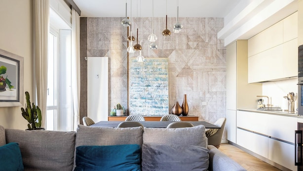 Een moderne woonkamer met een grijze bank, kussens in teal-tinten, en een eethoek met een wand met geometrische patronen en een groot abstract schilderij.