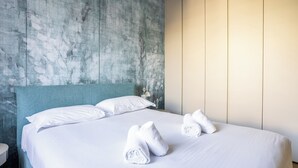 Een opgemaakt bed met witte lakens en handdoeken, een blauwgroen hoofdbord en een gestructureerde muur erachter.