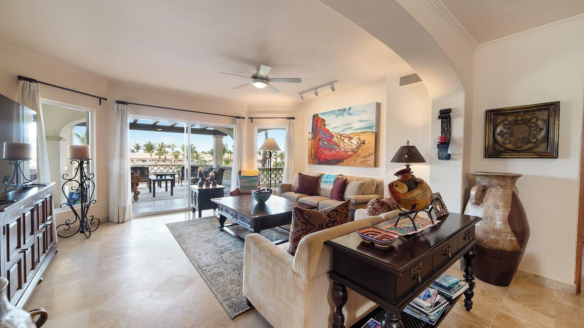 A spacious living area with a sofa, coffee table, and decorative items. There is a balcony with a view of palm trees.