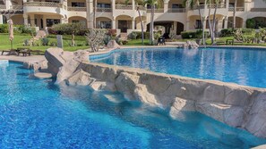 Una piscina con borde rocoso, rodeada por un edificio con varios balcones y un jardín cuidado.