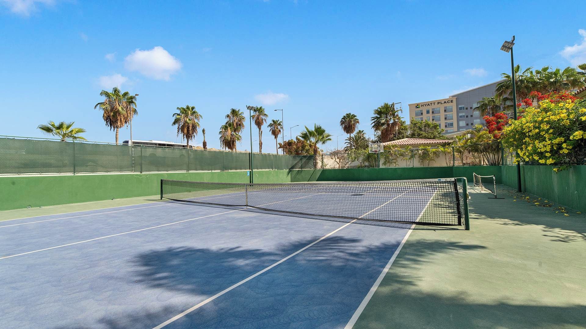 Un court de tennis avec un filet, entouré d’une clôture verte, de palmiers et d’un bâtiment sur lequel on peut lire l’inscription « HYATT PLACE ».